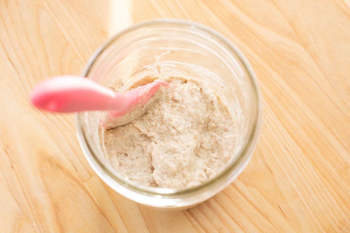 Mixing sourdough starter ingredients with a pink spatula in a glass jar.