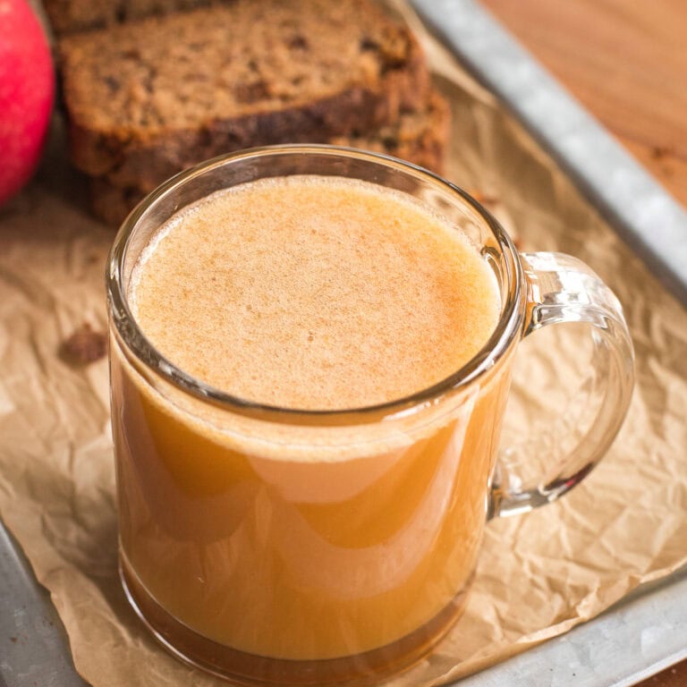 Glass mug of fresh apple cider on a tray with apple and slices of bread in the background.