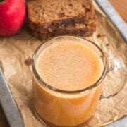 Glass mug of apple cider on a tray with apple and sliced bread behind it.