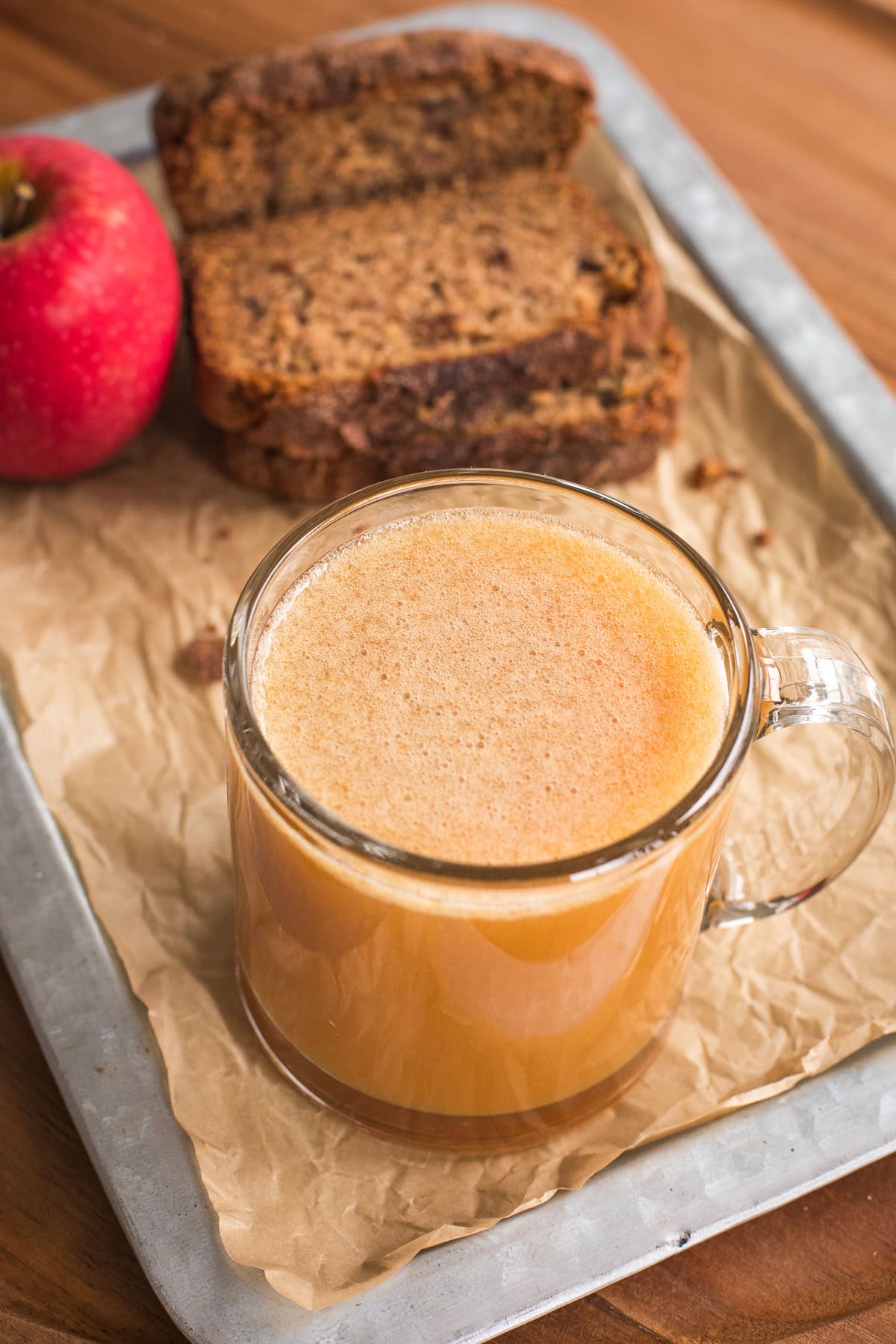 Glass mug of apple cider on a tray with apple and sliced bread behind it.