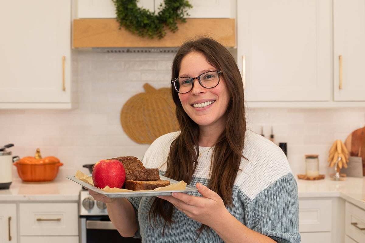 Smiling woman holding a tray with apple cinnamon bread and a red apple in a kitchen.