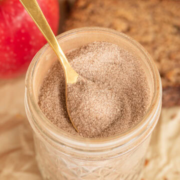 Close up of Glass jar of cinnamon sugar with a gold spoon, apple, and sliced bread in the background.