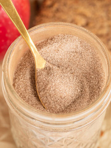 Close up of Glass jar of cinnamon sugar with a gold spoon, apple, and sliced bread in the background.