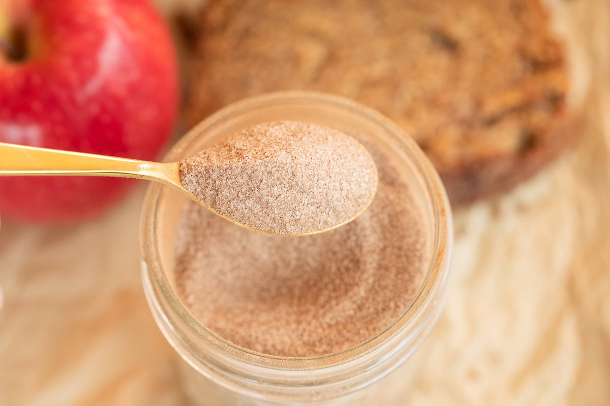 Spoonful of cinnamon sugar being held over a jar with apple and bread behind it.