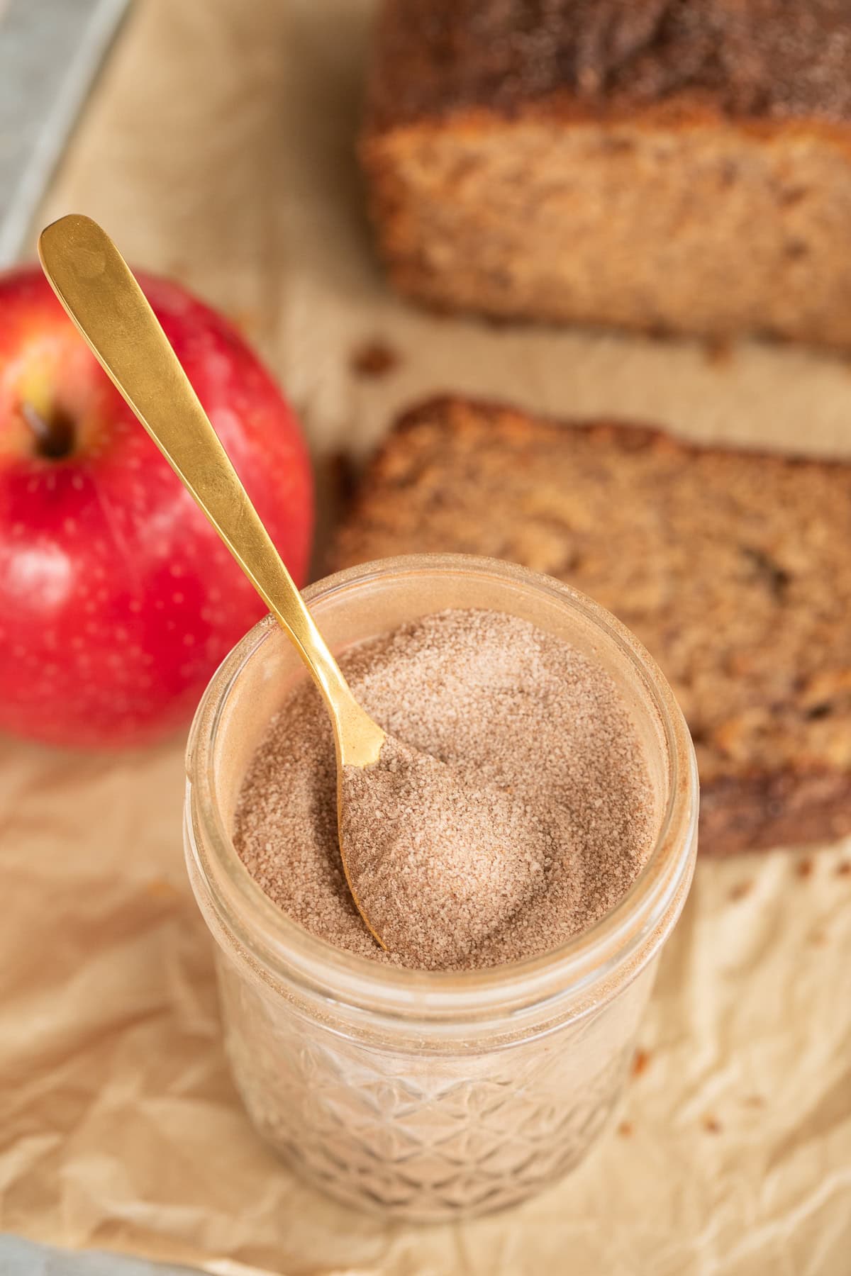 Glass jar of cinnamon sugar with a gold spoon, apple, and sliced bread in the background.