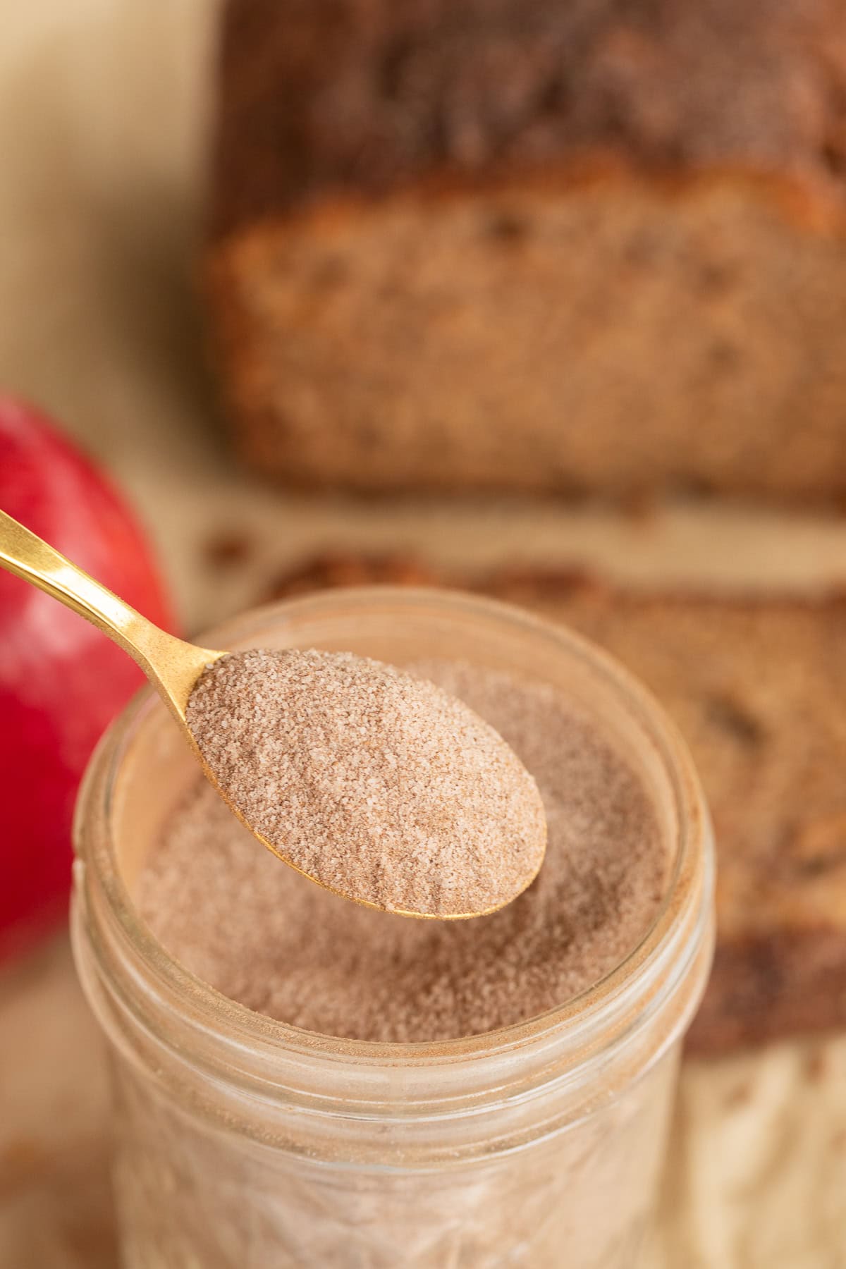 Spoonful of cinnamon sugar held over a jar with apple and bread behind it.