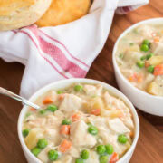 Two bowls of creamy slow cooker chicken pot pie with chicken, carrots, peas, and potatoes, served with a basket of golden biscuits.