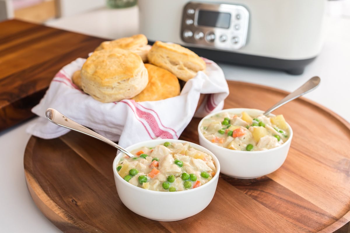 Slow cooker chicken pot pie served in two white bowls with biscuits in a towel-lined basket, GreenPan Elite slow cooker in background.