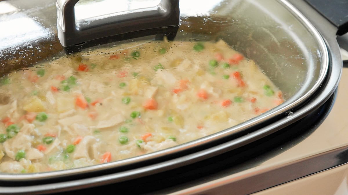 Slow cooker chicken pot pie from scratch simmering with peas, carrots, and potatoes visible through glass lid.