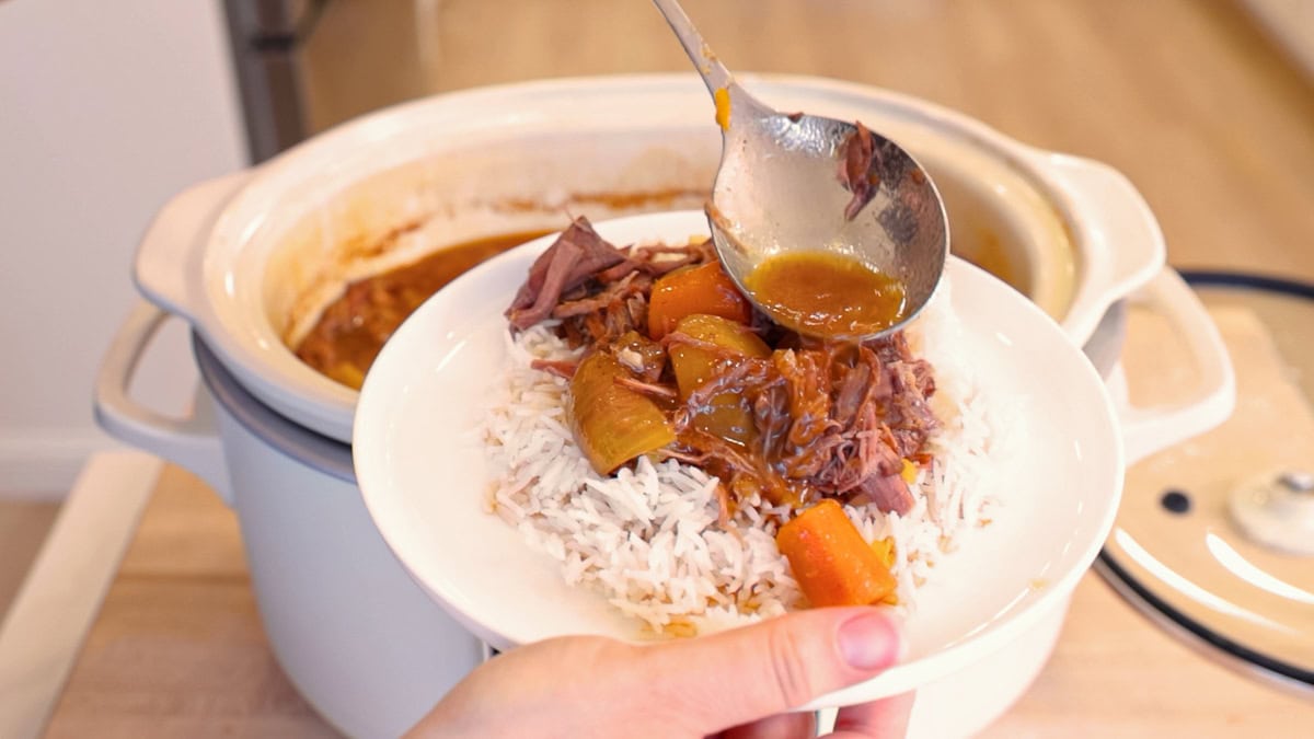 Hand holding a white plate of slow cooker pot roast with shredded beef, potatoes, carrots, and onions over rice while ladling sauce on top.