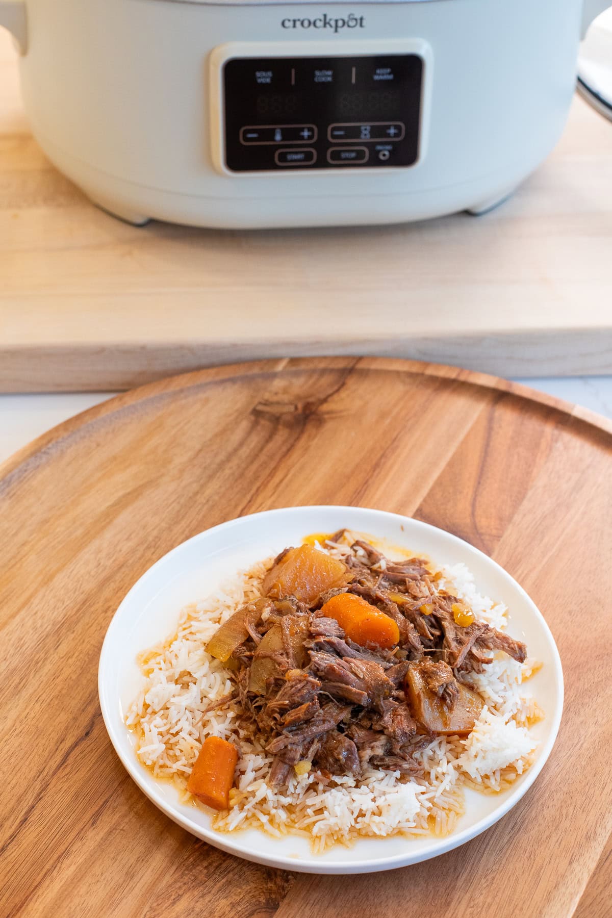Slow cooker pot roast on a plate with carrots and potatoes, Crock-Pot in the background.