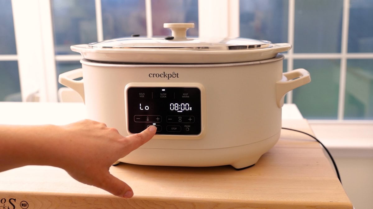 Hand pressing the start button on a Crock-Pot slow cooker set to low for 8 hours.