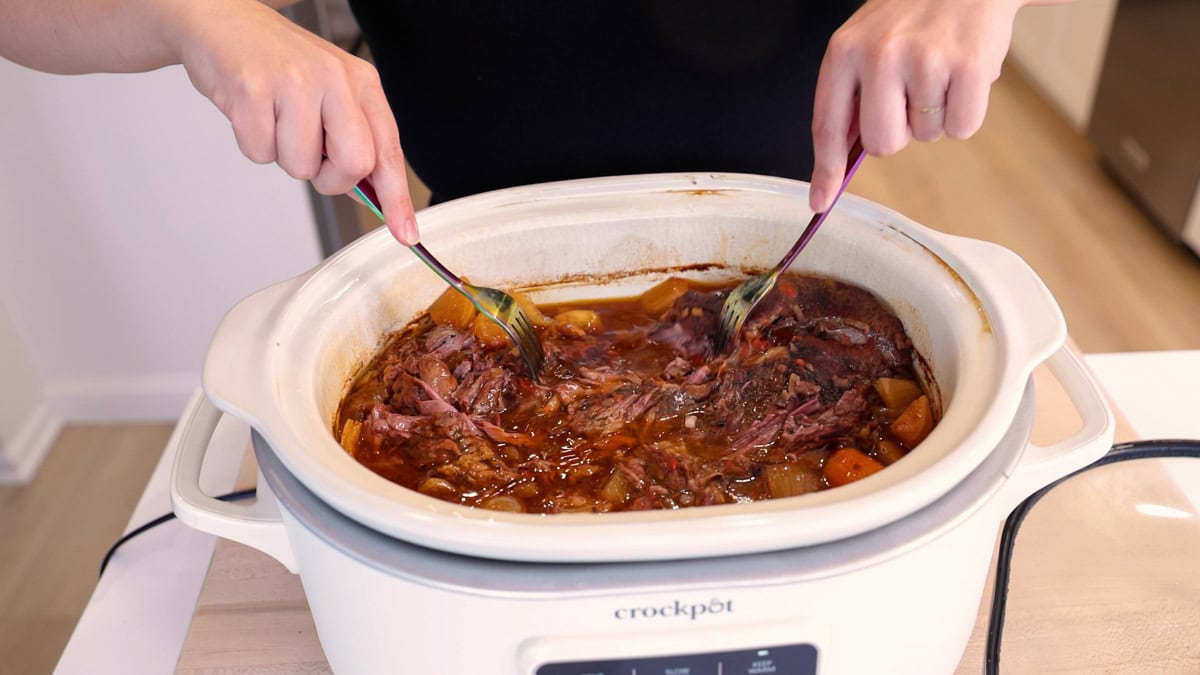 Shredding tender chuck roast with two forks inside a Crock-Pot slow cooker.