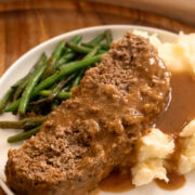 Close up of sliced meatloaf with brown gravy over mashed potatoes with green beans on a plate.