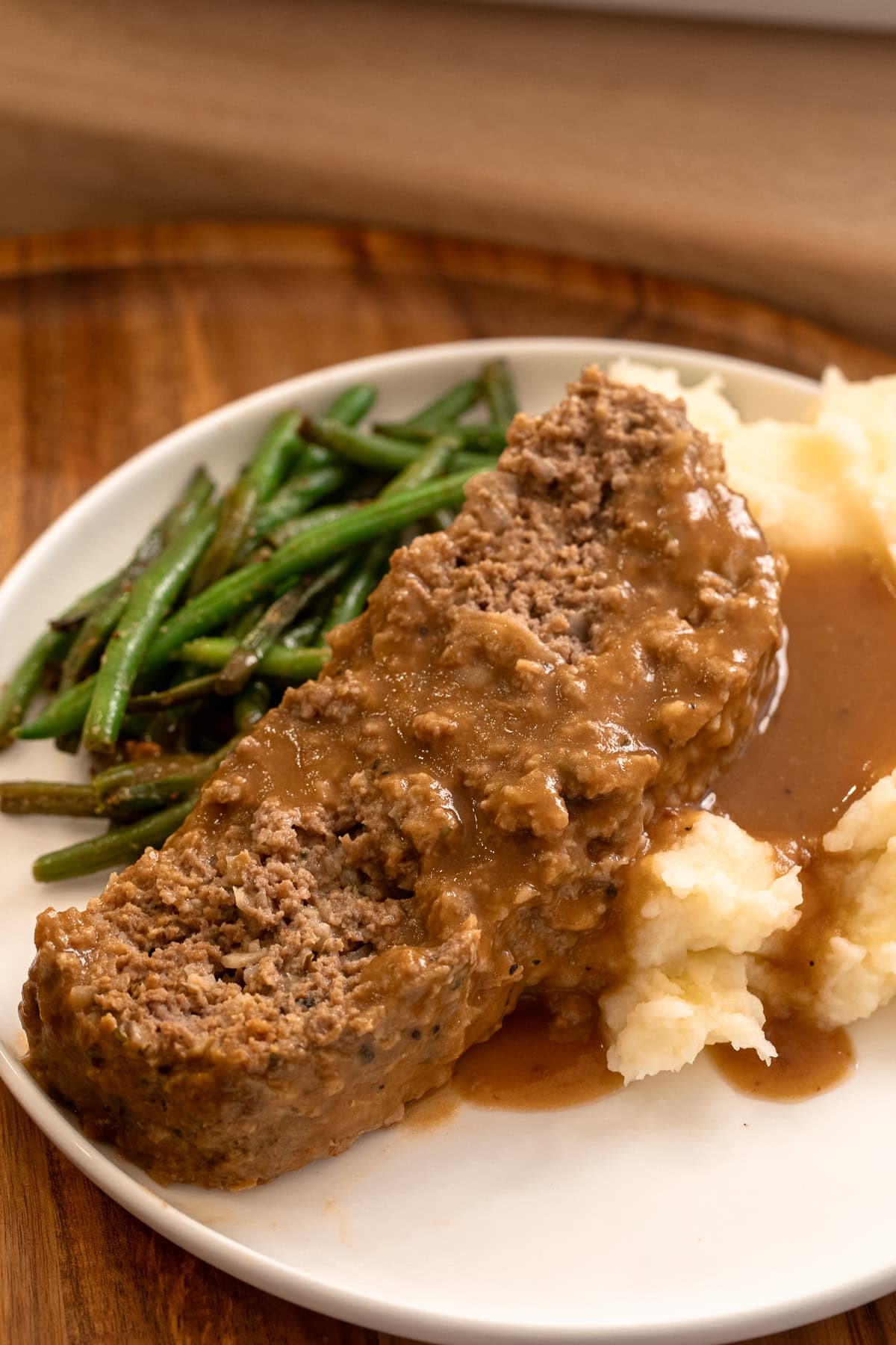 Close up of sliced meatloaf with brown gravy over mashed potatoes with green beans on a plate.