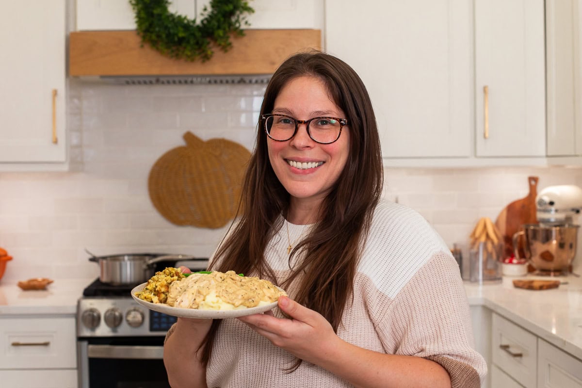 Woman smiling and holding a plate of slow cooker chicken and gravy with sides in a bright kitchen.