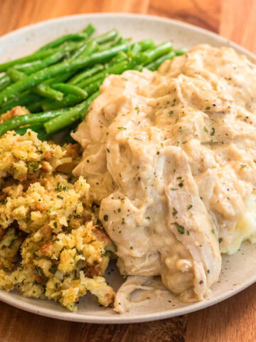 Slow cooker chicken and gravy recipe served over mashed potatoes on a plate with garlic string beans and stuffing.
