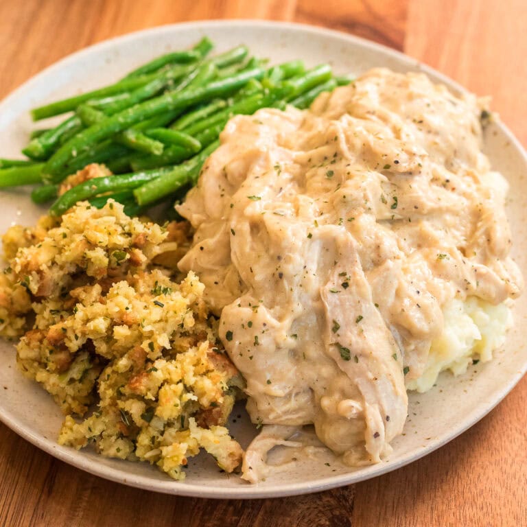 Slow cooker chicken and gravy recipe served over mashed potatoes on a plate with garlic string beans and stuffing.