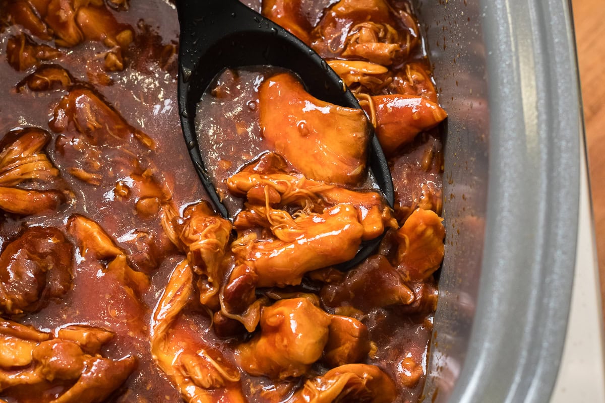 slow cooker filled with honey garlic BBQ chicken thighs falling apart in a glossy, rich sauce with a serving spoon.