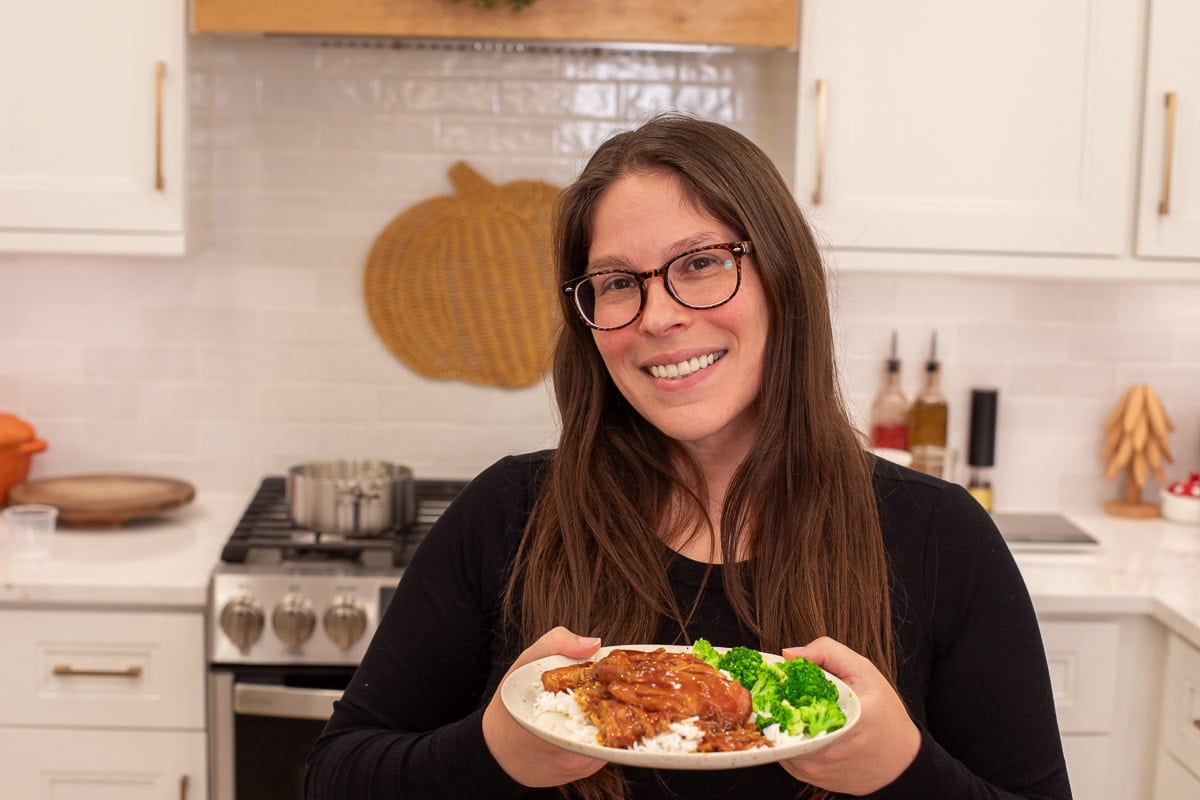 Woman in a bright kitchen holding a plate of honey garlic BBQ chicken with rice and broccoli, smiling at the camera.