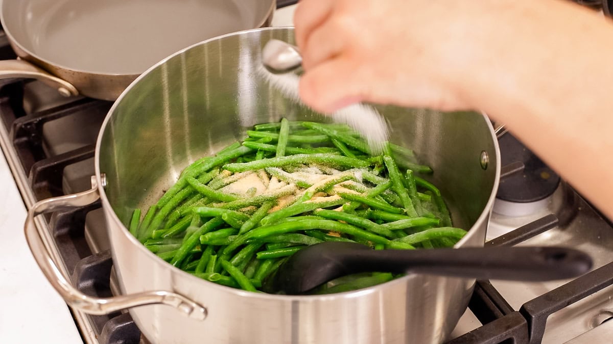Hand sprinkling seasoning over frozen green beans in a stainless steel pot on the stove.