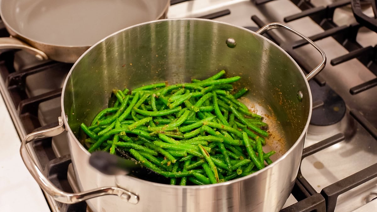 Frozen green beans cooking in a stainless steel pot with seasonings starting to coat them.