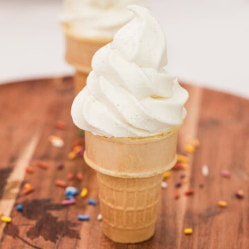 Two vanilla soft serve ice cream cones on a wooden cutting board with sprinkles.