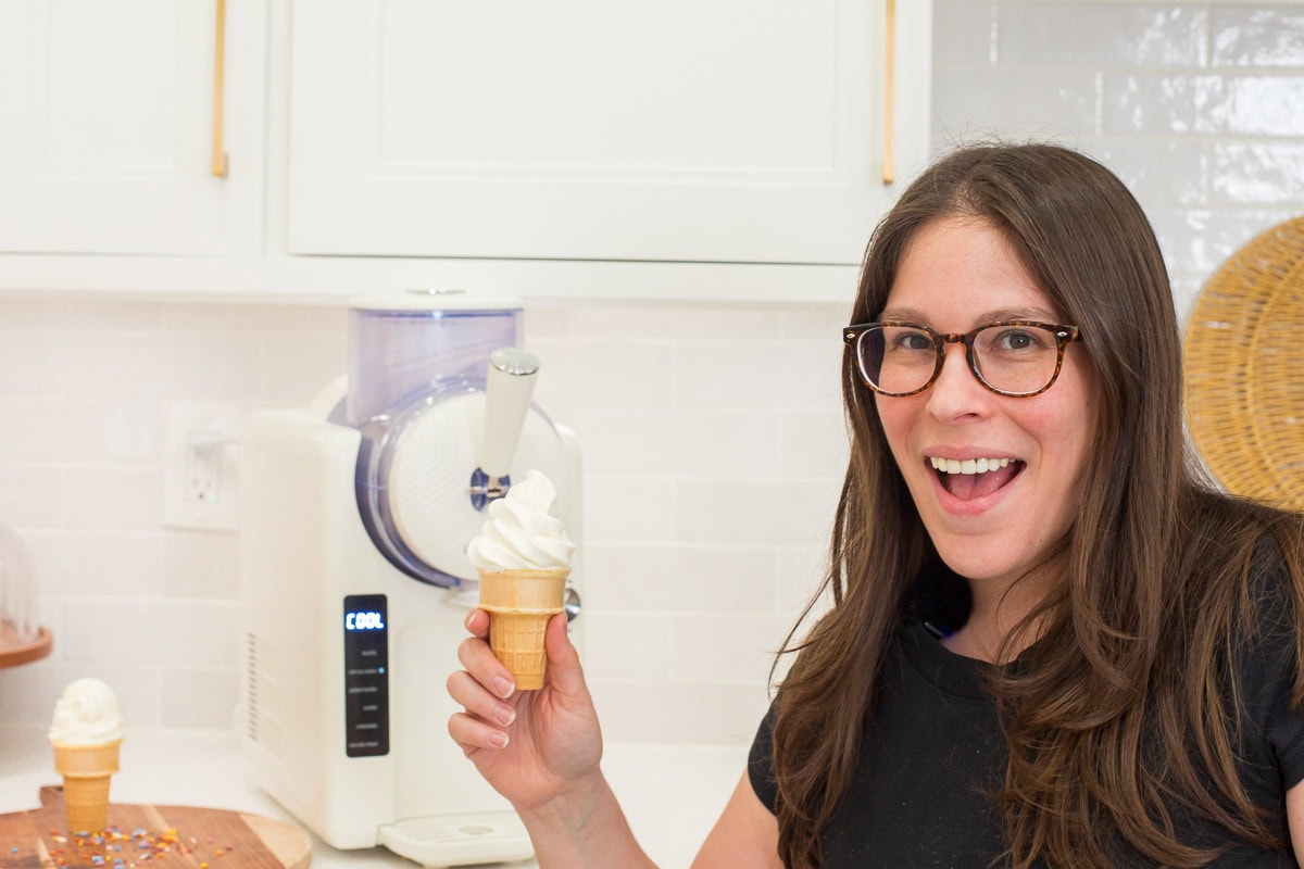 Courtney of Courtney's Sweets holding a vanilla soft serve ice cream cone in her kitchen next to the GreenPan Frost frozen ice cream and frozen drink maker.