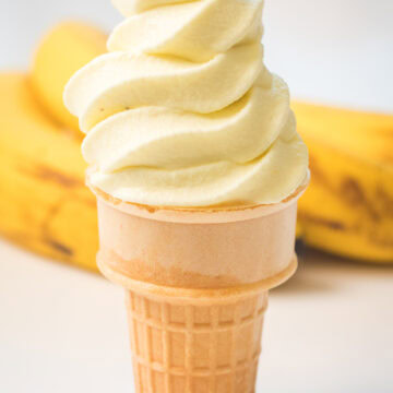 Banana soft serve in an ice cream cone with ripe bananas behind it.
