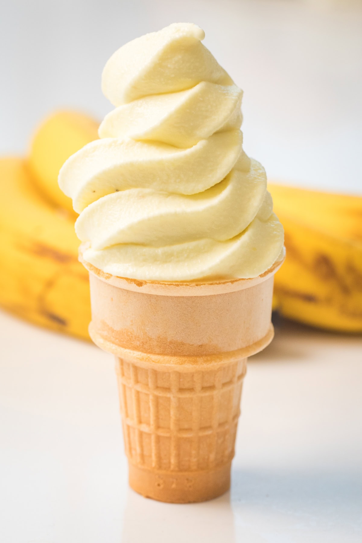 Banana soft serve in an ice cream cone with ripe bananas behind it.