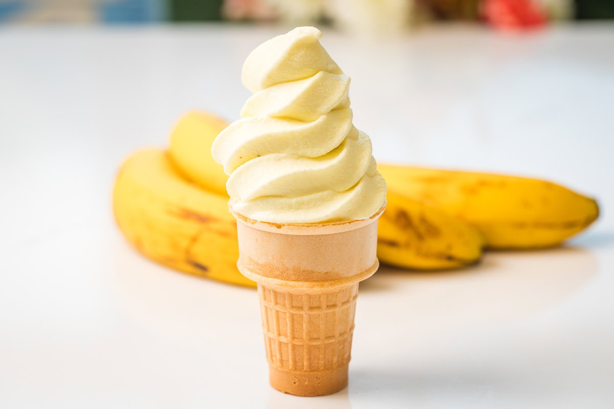 close up of banana soft serve in an ice cream cone with ripe bananas behind it.