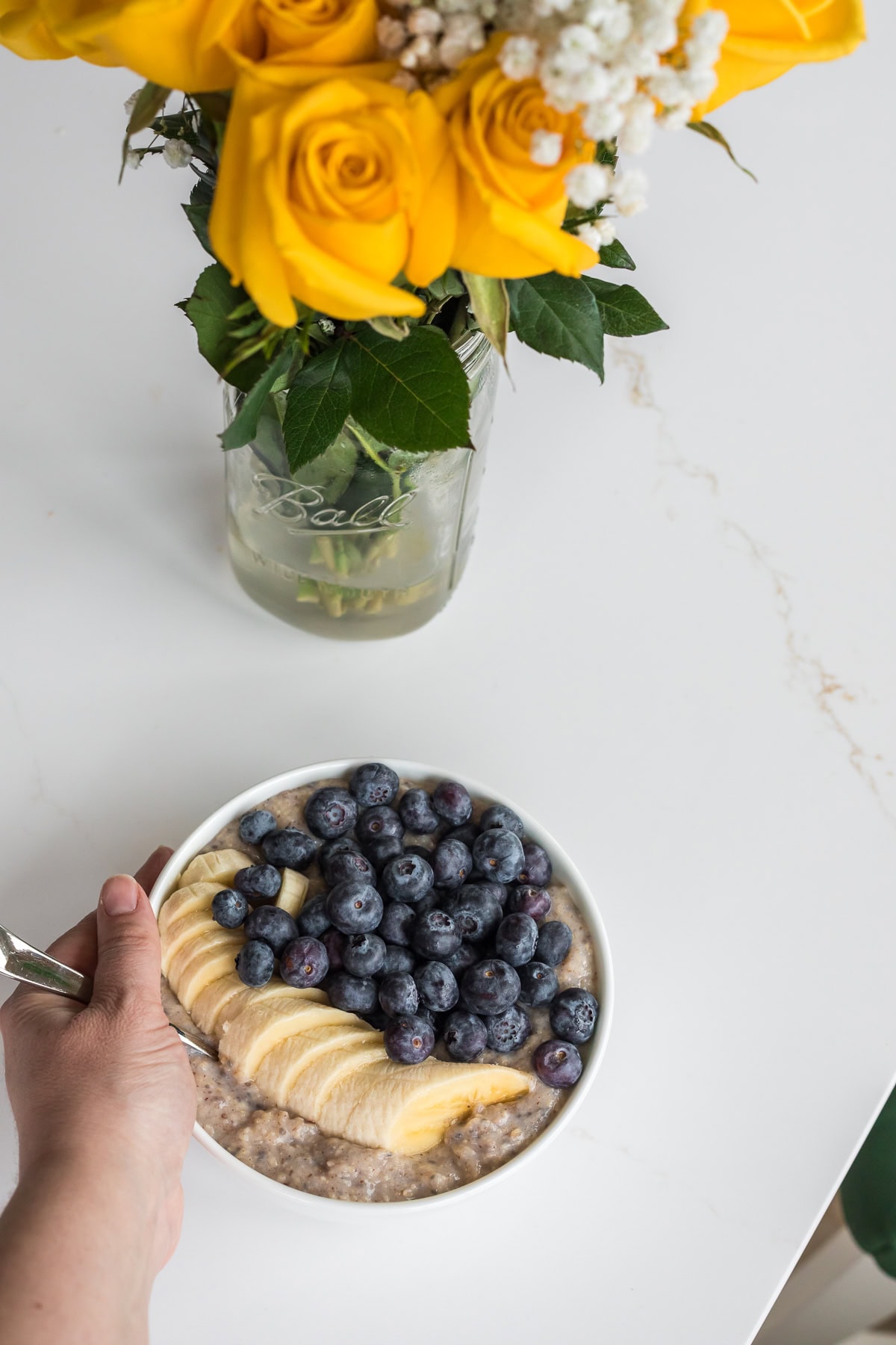 Overhead view of a hand holding a white bowl of oat bran topped with banana and blueberries on a white countertop, with a vase of yellow roses in the background.