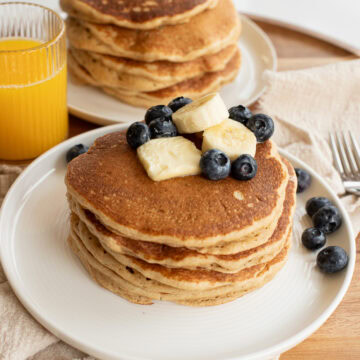 fresh milled flour pancakes on a white plate topped with a pat of butter, banana slices and blueberries.