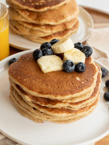 fresh milled flour pancakes on a white plate topped with a pat of butter, banana slices and blueberries.