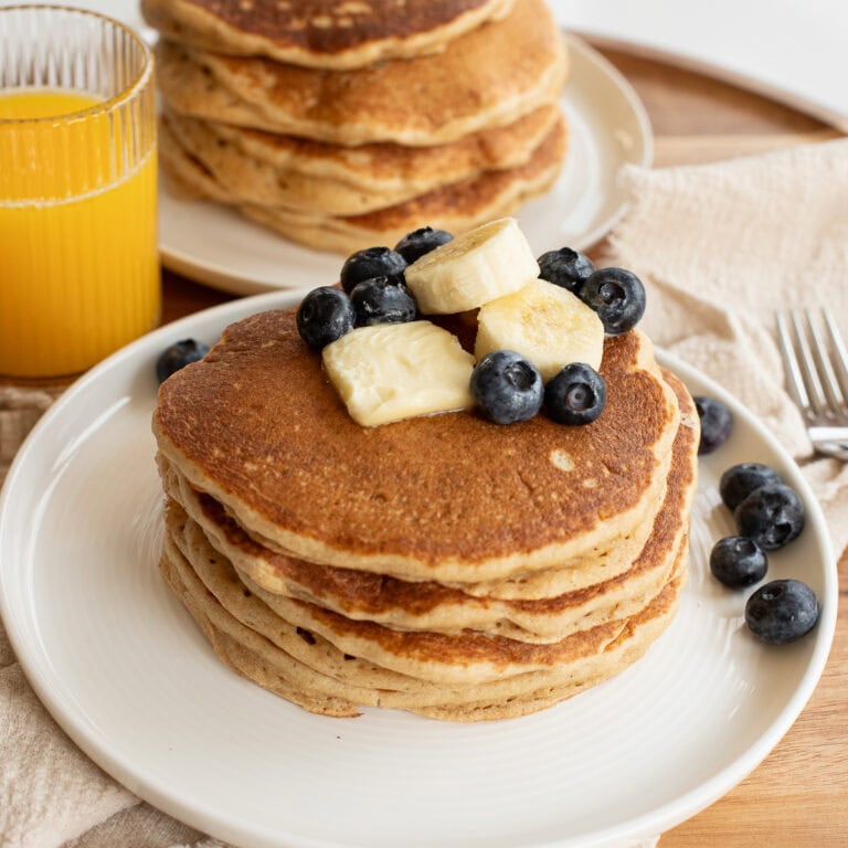 fresh milled flour pancakes on a white plate topped with a pat of butter, banana slices and blueberries.
