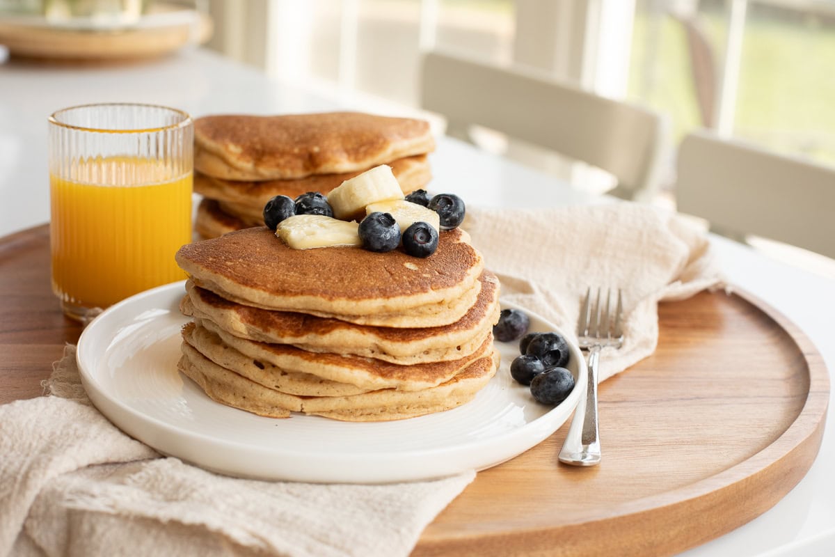 stacked fresh milled flour pancakes on a white plate topped with a pat of butter, banana slices and blueberries.