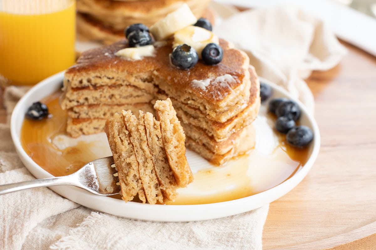 Fork with bite of pancakes with a stack of fresh milled flour pancakes on a white plate topped with a pat of butter, banana slices and blueberries.