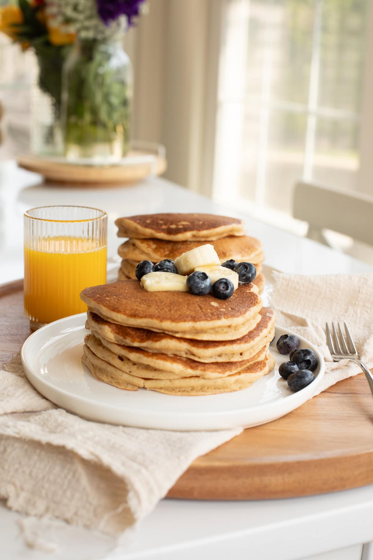 fresh milled flour pancakes on a white plate topped with a pat of butter, banana slices and blueberries on counter with flowers and windows in background.