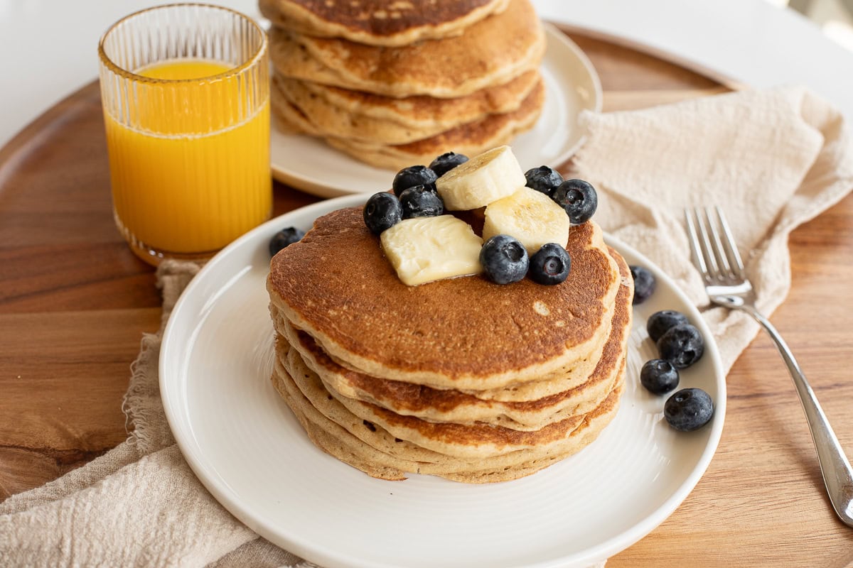 homemade fresh milled flour pancakes on a white plate topped with a pat of butter, banana slices and blueberries.