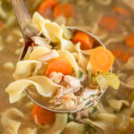 Ladle full of slow cooker chicken noodle soup with chicken, carrots, celery, and egg noodles over a bowl of soup.