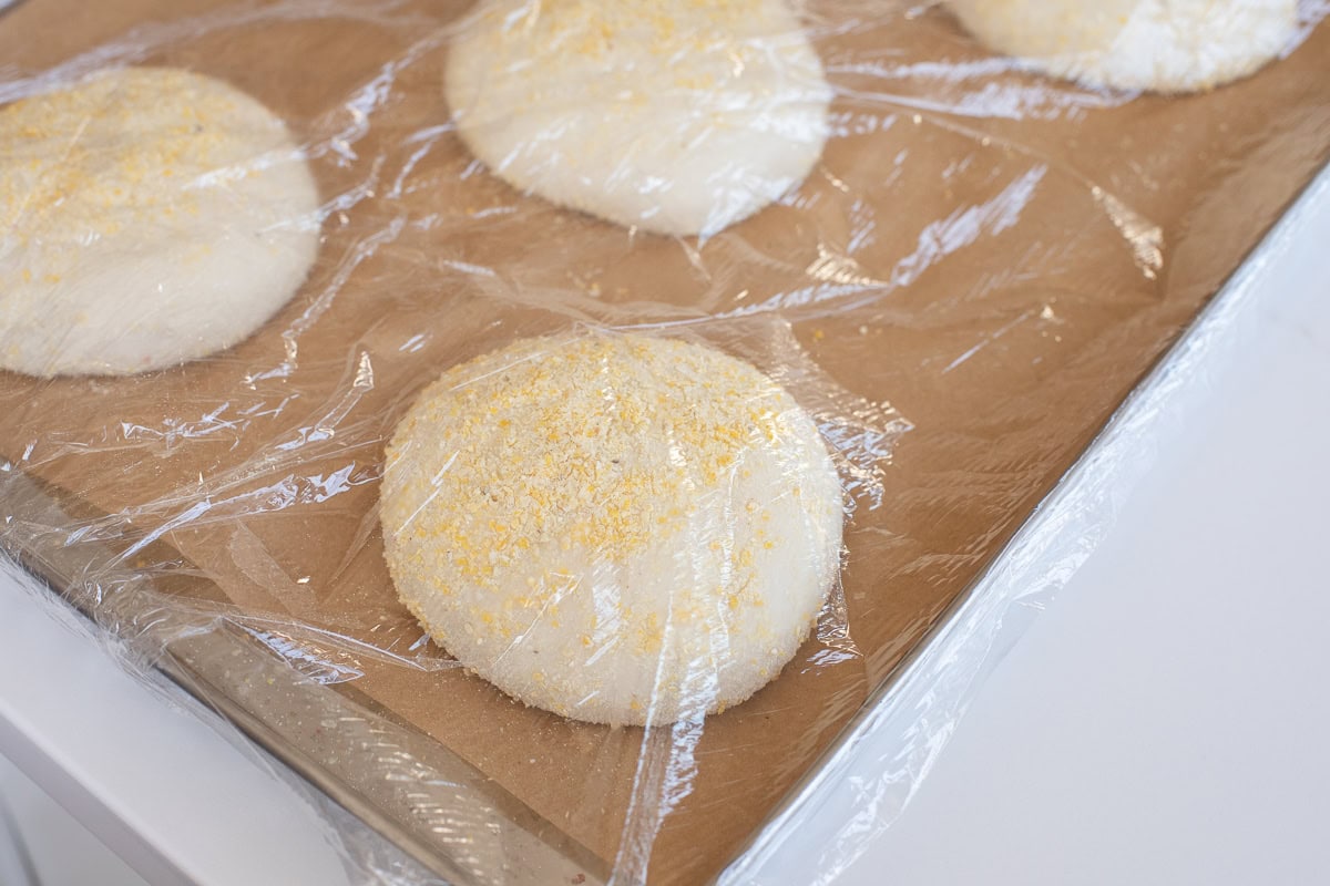 proofed english muffins under plastic wrap with cornmeal on them on a parchment lined baking sheet.