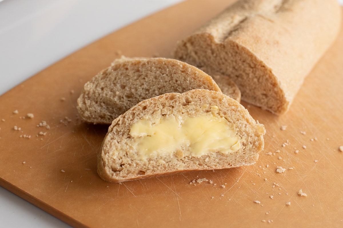 Fresh milled flour french bread on a cutting board sliced and whole, front slice has butter spread on it.
