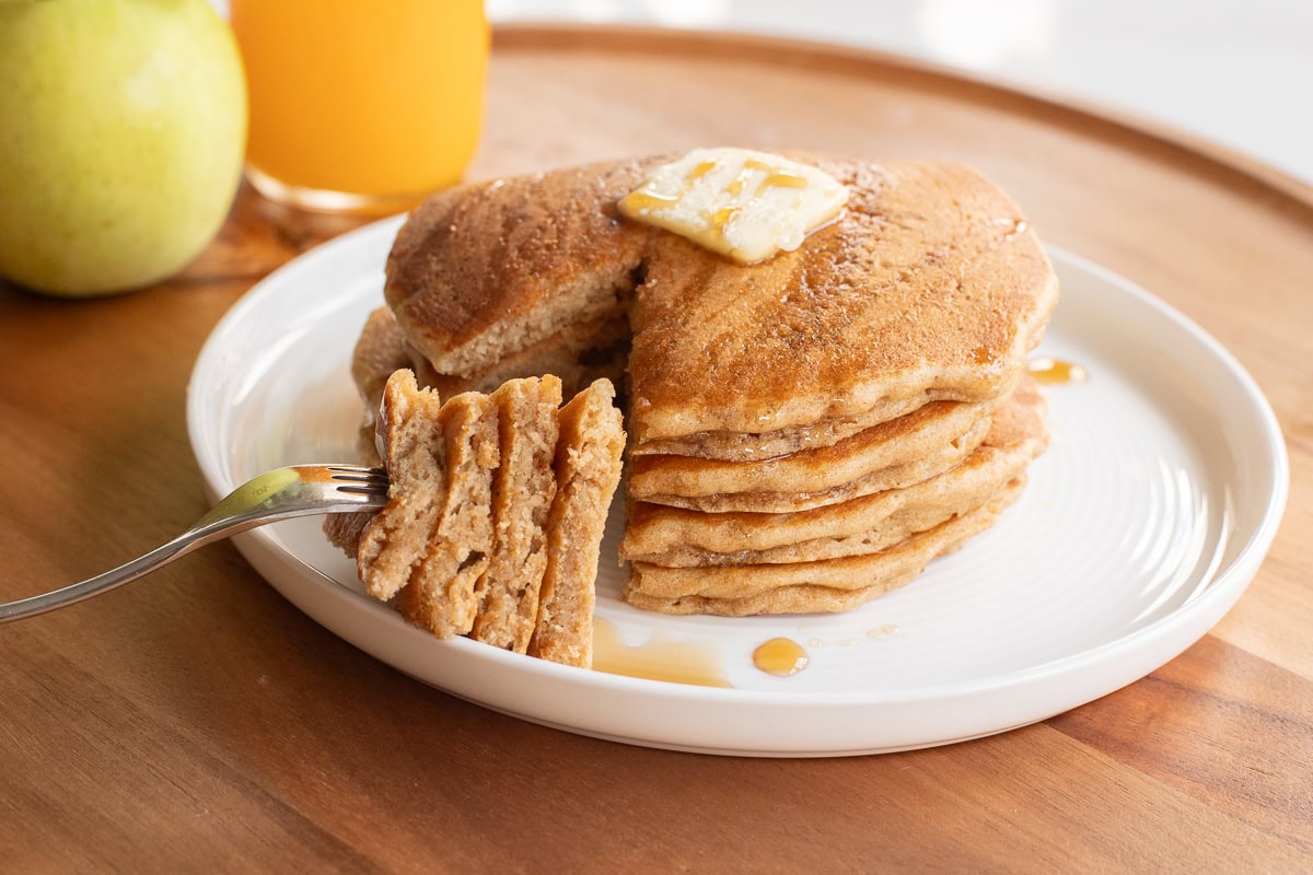 Stack of Einkorn pancakes with a pat of butter and a drizzle of maple syrup and a fork holding some to show the texture.