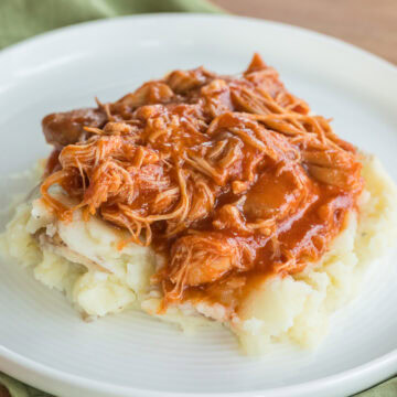 Homemade Slow Cooker BBQ Chicken Thighs over mashed potatoes on a white plate with a green napkin under it.