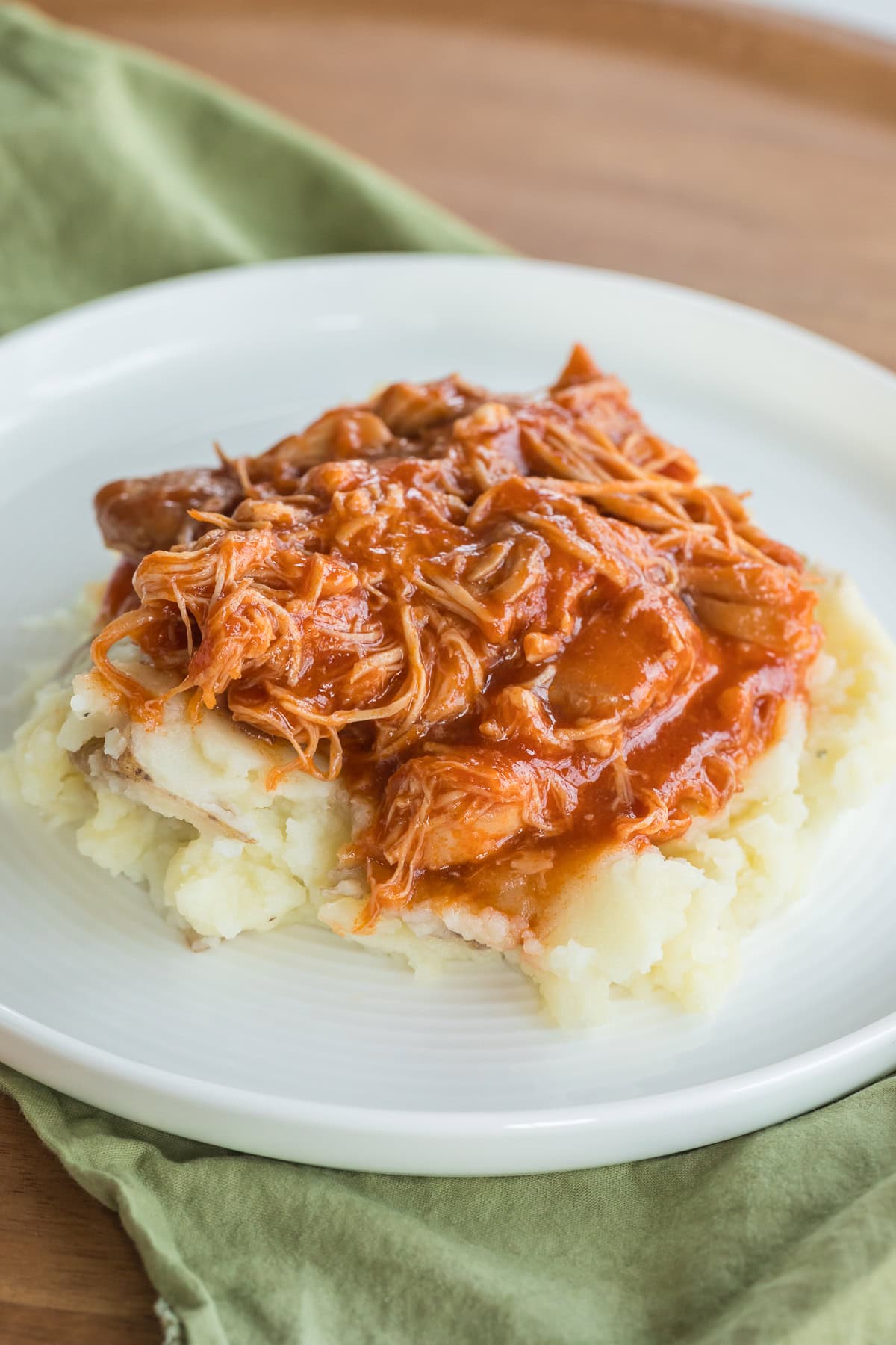 Homemade Slow Cooker BBQ Chicken Thighs over mashed potatoes on a white plate with a green napkin under it.
