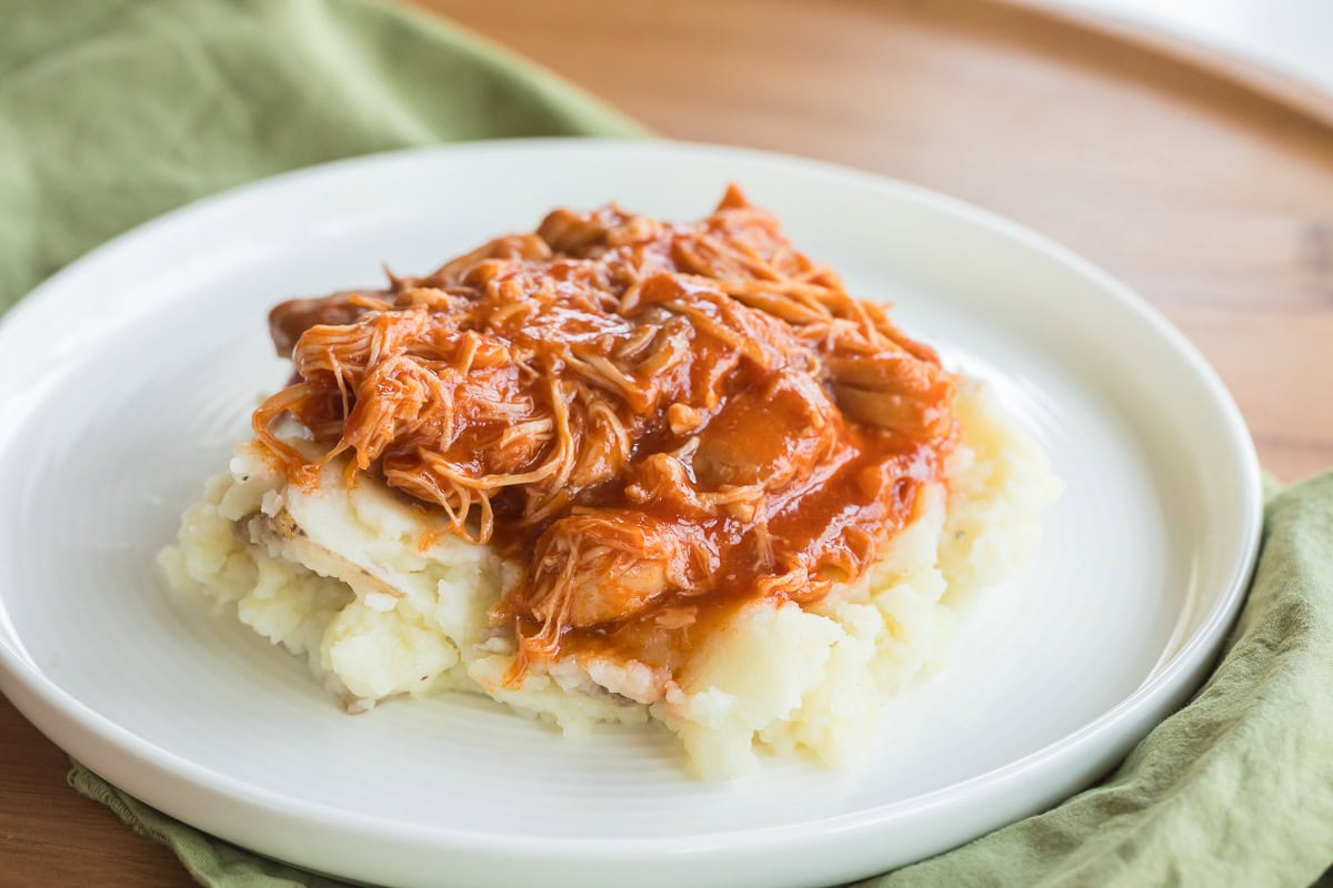 Slow Cooker BBQ Chicken Thighs over mashed potatoes on a white plate with a green napkin under it.